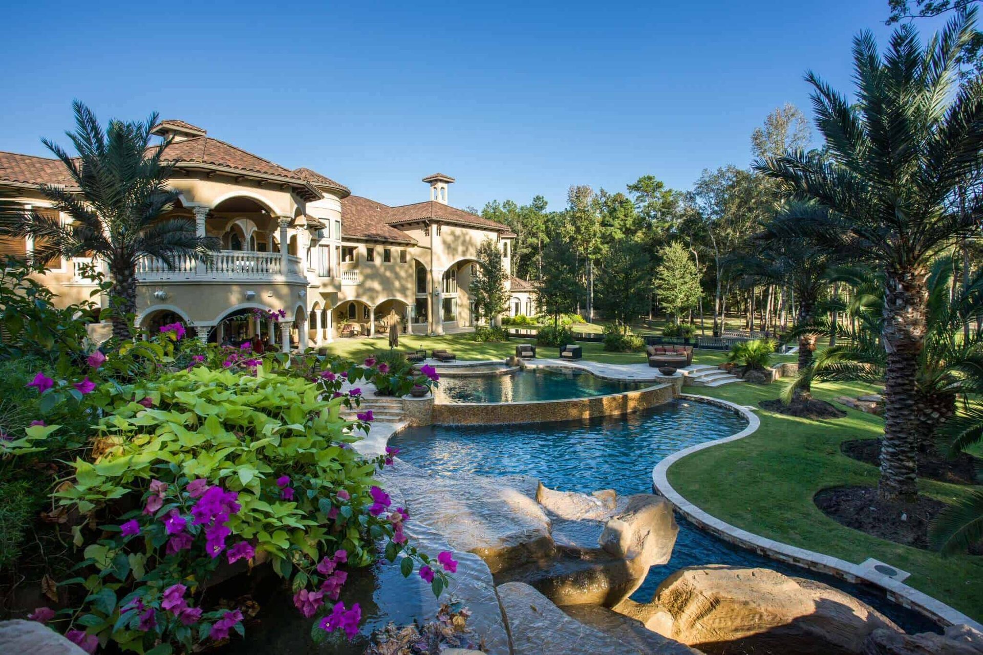 A winding backyard pool with palm trees, flowering landscaping, and stone waterfalls